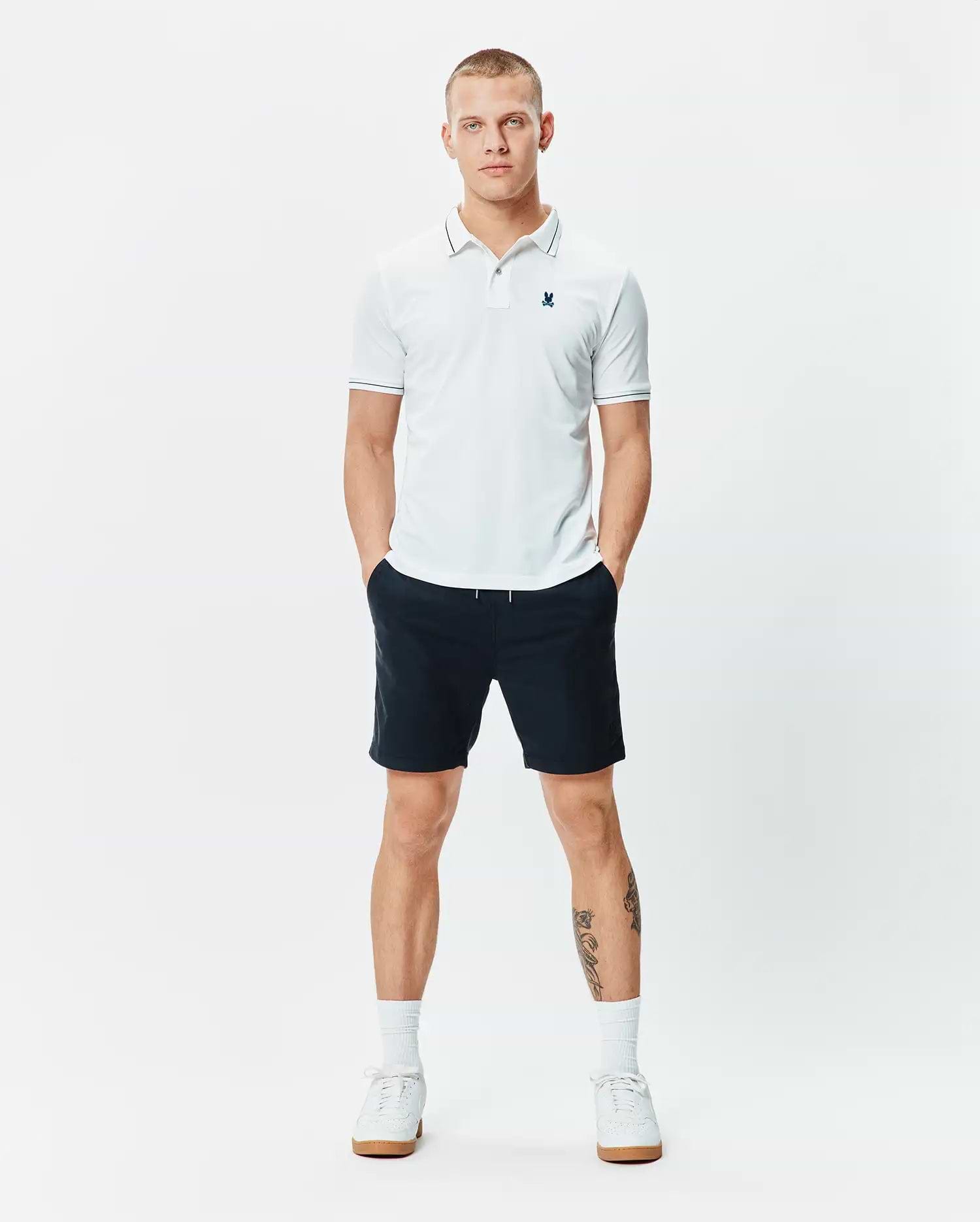 Mens Edwin Sport Polo, a classic fit white pique polo with navy tipping on the collar and cuffs, featuring a small embroidered teal Psycho Bunny skull logo on the left chest.