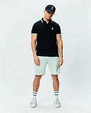 Mens Holden Pique Polo, black, classic fit, white striped collar/cuffs, small embroidered white Psycho Bunny skull logo on left chest. Light green shorts, black Psycho Bunny trucker cap.