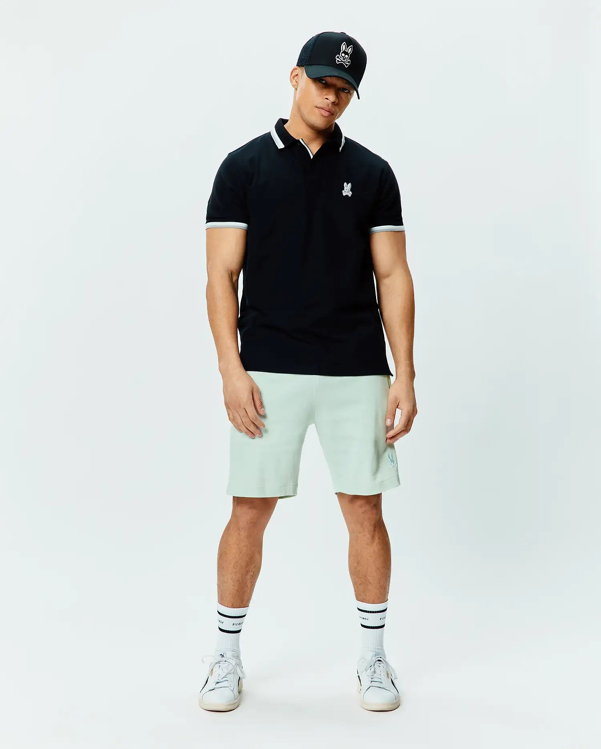 Mens Holden Pique Polo, black, classic fit, white striped collar/cuffs, small embroidered white Psycho Bunny skull logo on left chest. Light green shorts, black Psycho Bunny trucker cap.