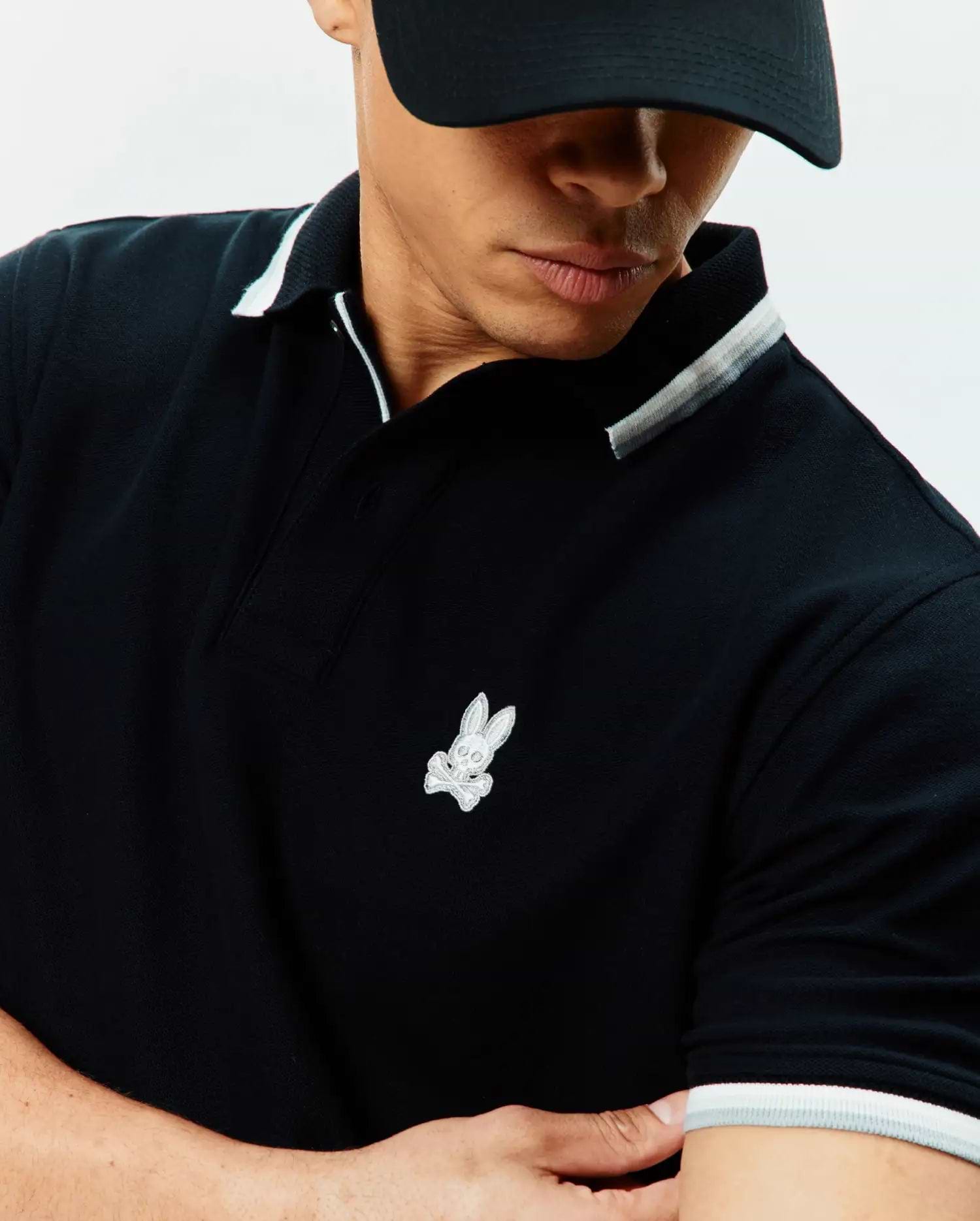 Mens Holden Pique Polo in black, featuring a classic fit, pique fabric, ribbed collar and cuffs with white and light grey tipping, and a small embroidered white Psycho Bunny skull logo on the left chest.