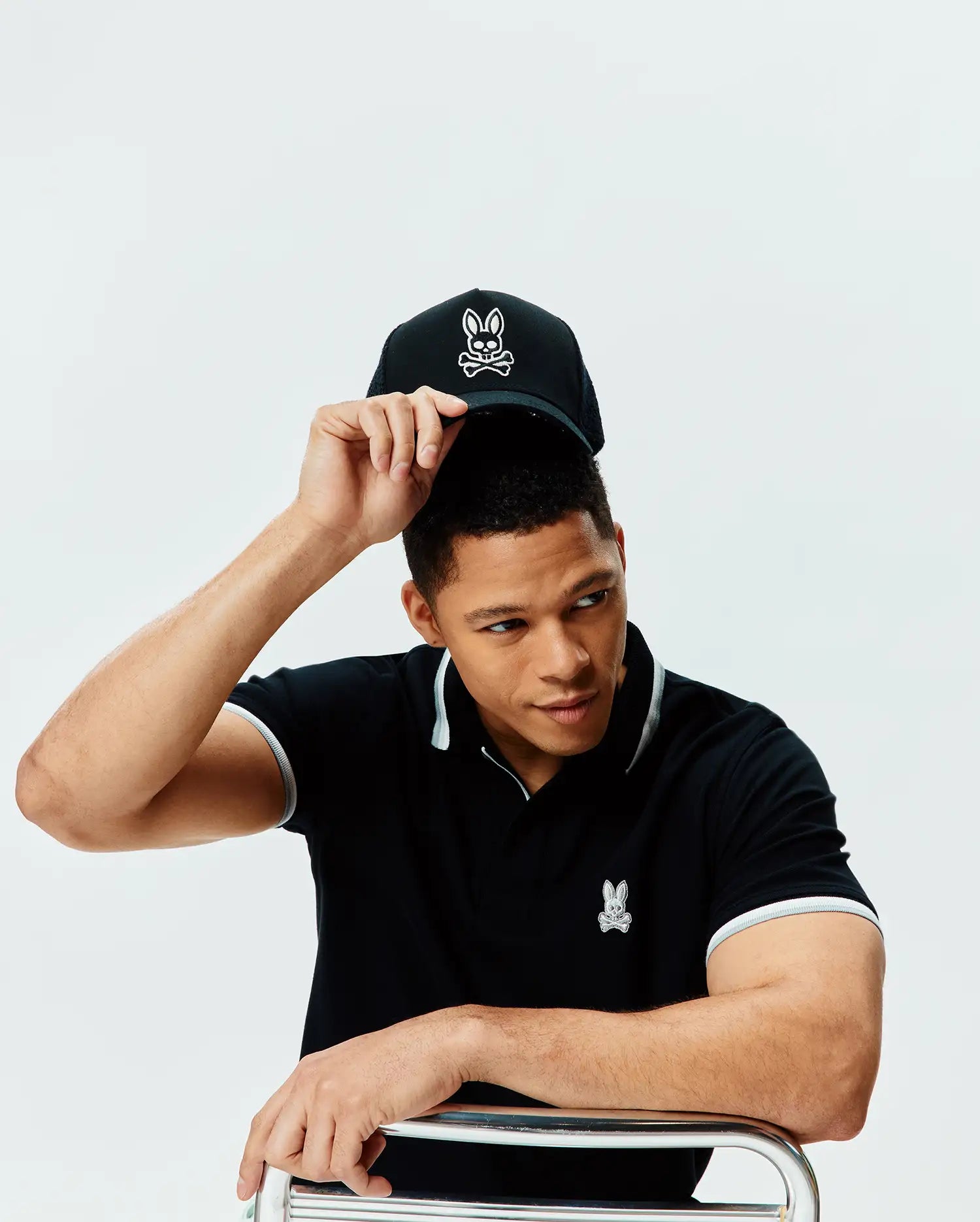 Mens Holden Pique Polo, black classic fit pique with light grey collar/cuff tipping, small embroidered white Psycho Bunny skull logo on chest. Black cap with large white bunny skull logo.