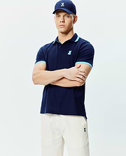 Mens Holden Pique Polo, navy pique, light blue contrast tipping on collar and cuffs, small embroidered Psycho Bunny skull logo on left chest. Cream shorts and navy cap also feature the logo.