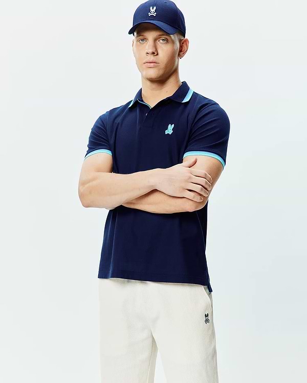 Mens Holden Pique Polo, navy pique, light blue contrast tipping on collar and cuffs, small embroidered Psycho Bunny skull logo on left chest. Cream shorts and navy cap also feature the logo.