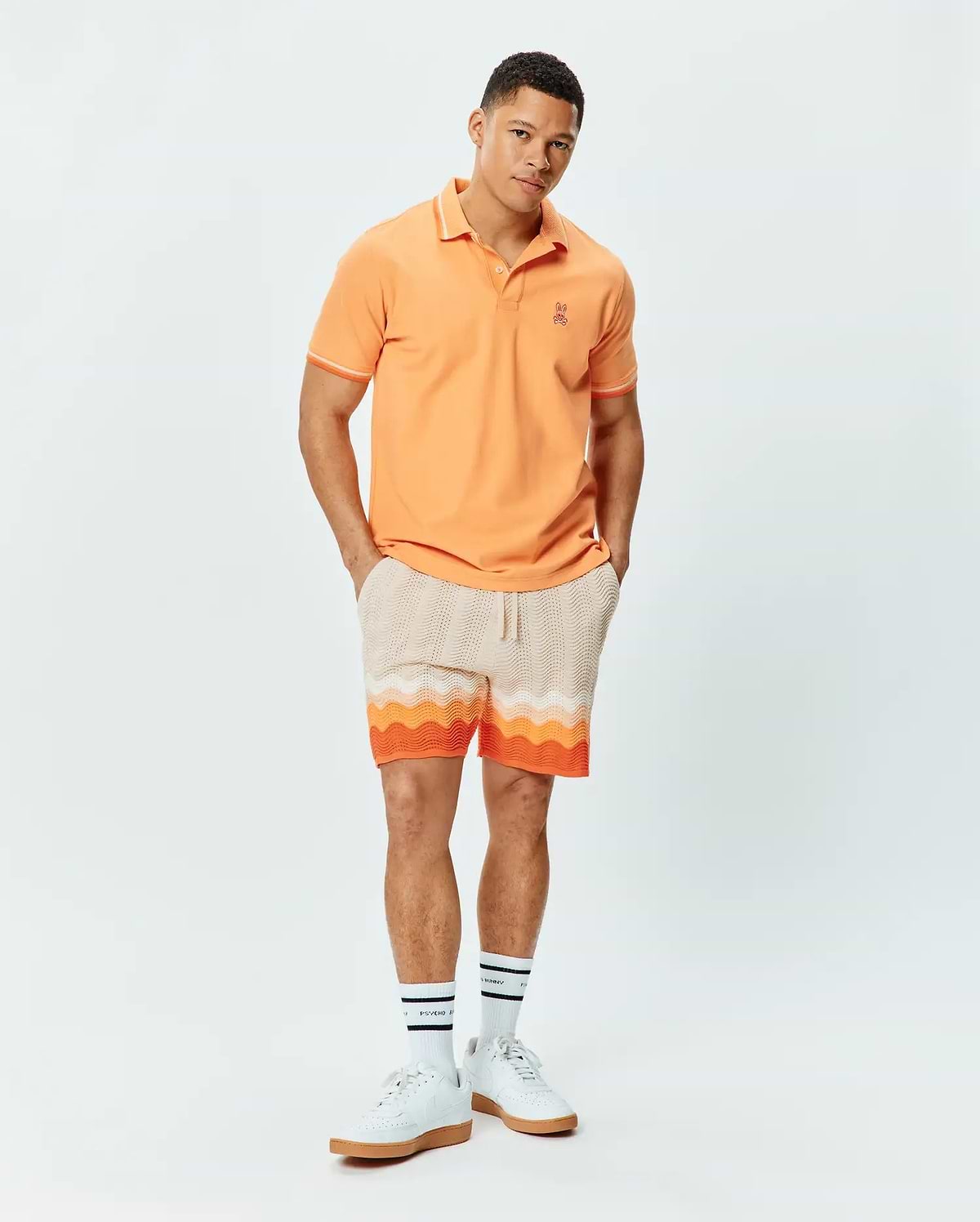 Mens Holden Pique Polo in peach with white trim, featuring a small embroidered Psycho Bunny skull logo on the left chest. Paired with cream knit shorts showcasing a vibrant orange and white wavy pattern.
