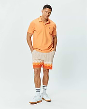 Mens Holden Pique Polo in peach with white trim, featuring a small embroidered Psycho Bunny skull logo on the left chest. Paired with cream knit shorts showcasing a vibrant orange and white wavy pattern.