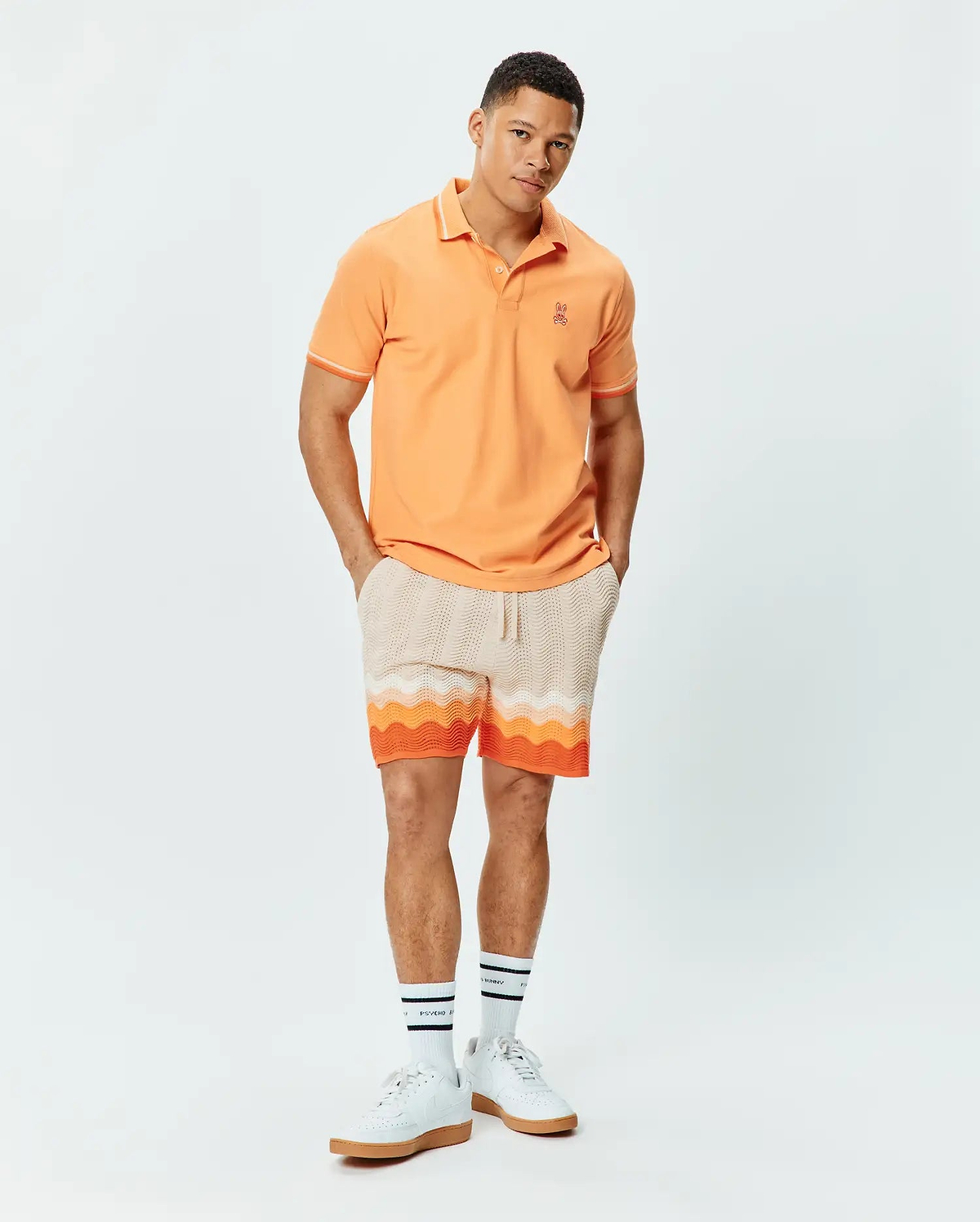 Mens Holden Pique Polo in peach with white trim, featuring a small embroidered Psycho Bunny skull logo on the left chest. Paired with cream knit shorts showcasing a vibrant orange and white wavy pattern.