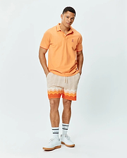 Mens Holden Pique Polo in peach with white trim, featuring a small embroidered Psycho Bunny skull logo on the left chest. Paired with cream knit shorts showcasing a vibrant orange and white wavy pattern.