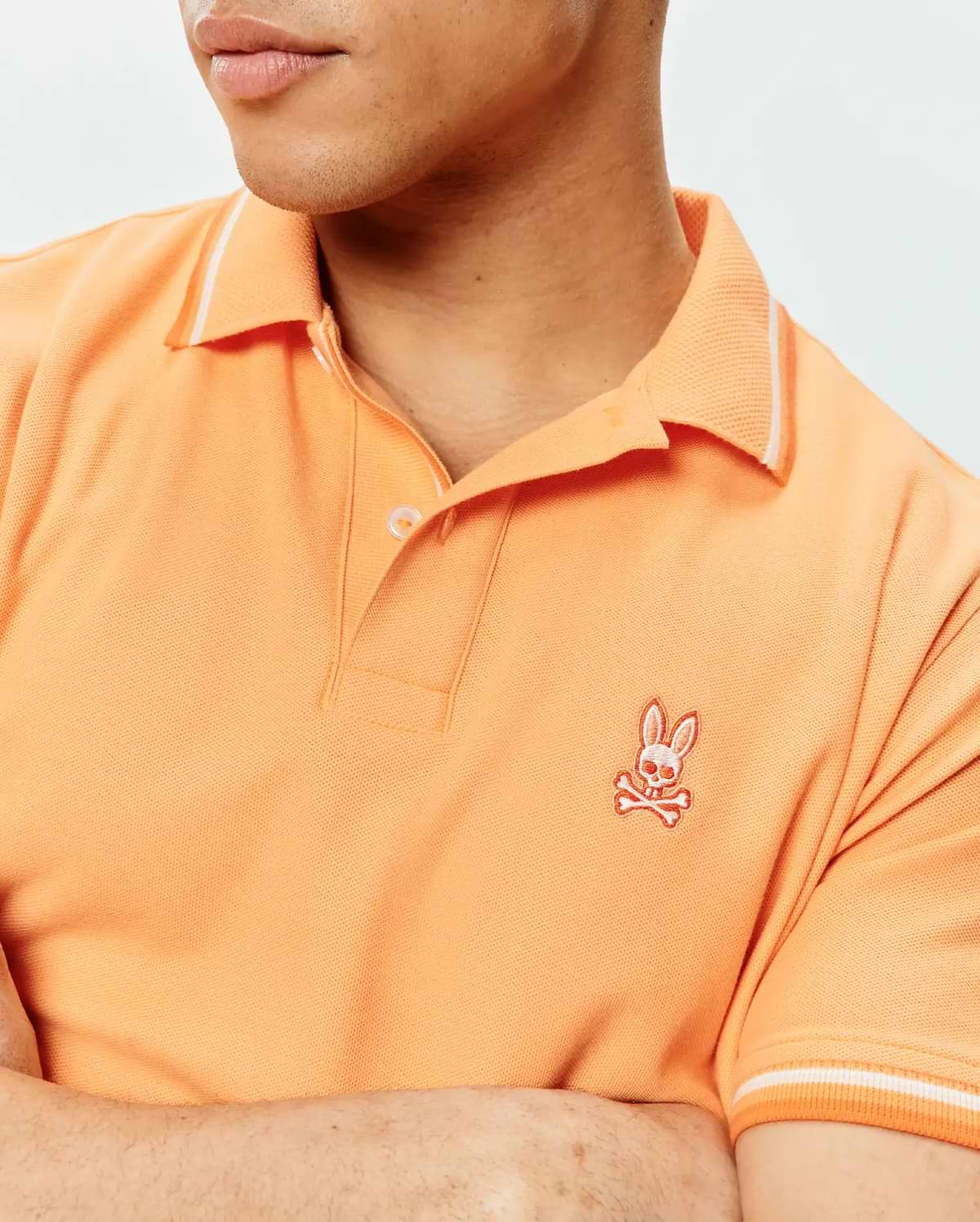 Mens Holden Pique Polo in vibrant orange, featuring a classic fit, pique knit texture, white tipped ribbed collar and cuffs, and an embroidered Psycho Bunny skull logo on the left chest.