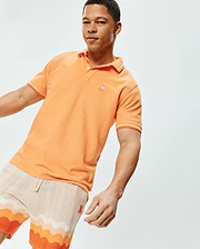 Mens Holden Pique Polo, a vibrant orange classic fit pique knit polo with white tipping on the ribbed collar and sleeve cuffs, featuring a small embroidered white Psycho Bunny skull logo on the left chest.
