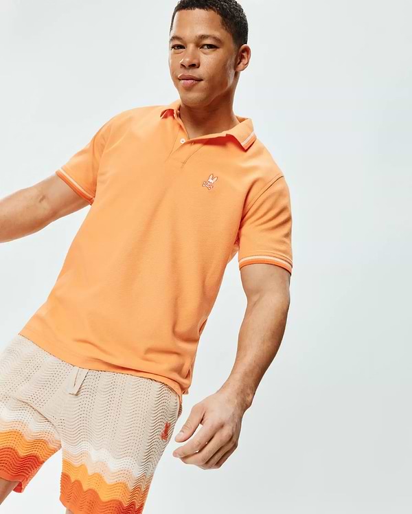 Mens Holden Pique Polo, a vibrant orange classic fit pique knit polo with white tipping on the ribbed collar and sleeve cuffs, featuring a small embroidered white Psycho Bunny skull logo on the left chest.