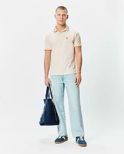 Mens Holden Pique Polo in cream, featuring a classic fit, pique texture, and orange tipping on the collar and sleeve cuffs, with a small embroidered orange Psycho Bunny skull logo on the left chest.