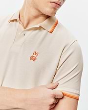 Mens Holden Pique Polo in beige pique knit, featuring a small orange embroidered Psycho Bunny skull logo on the left chest and orange striped collar and sleeve cuffs.
