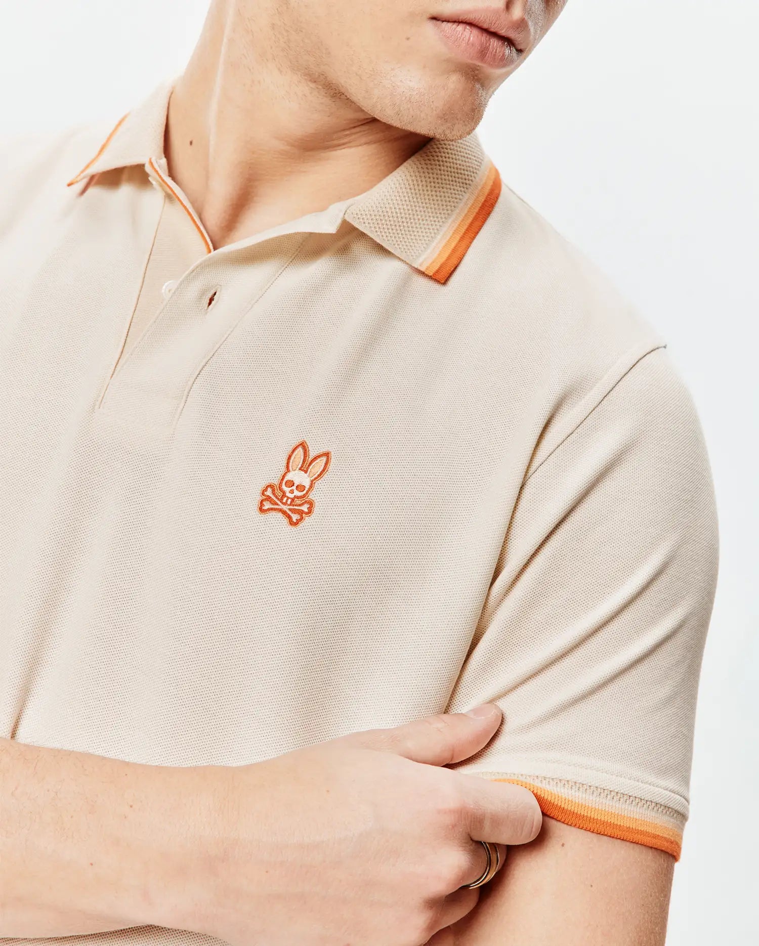 Mens Holden Pique Polo in beige pique knit, featuring a small orange embroidered Psycho Bunny skull logo on the left chest and orange striped collar and sleeve cuffs.