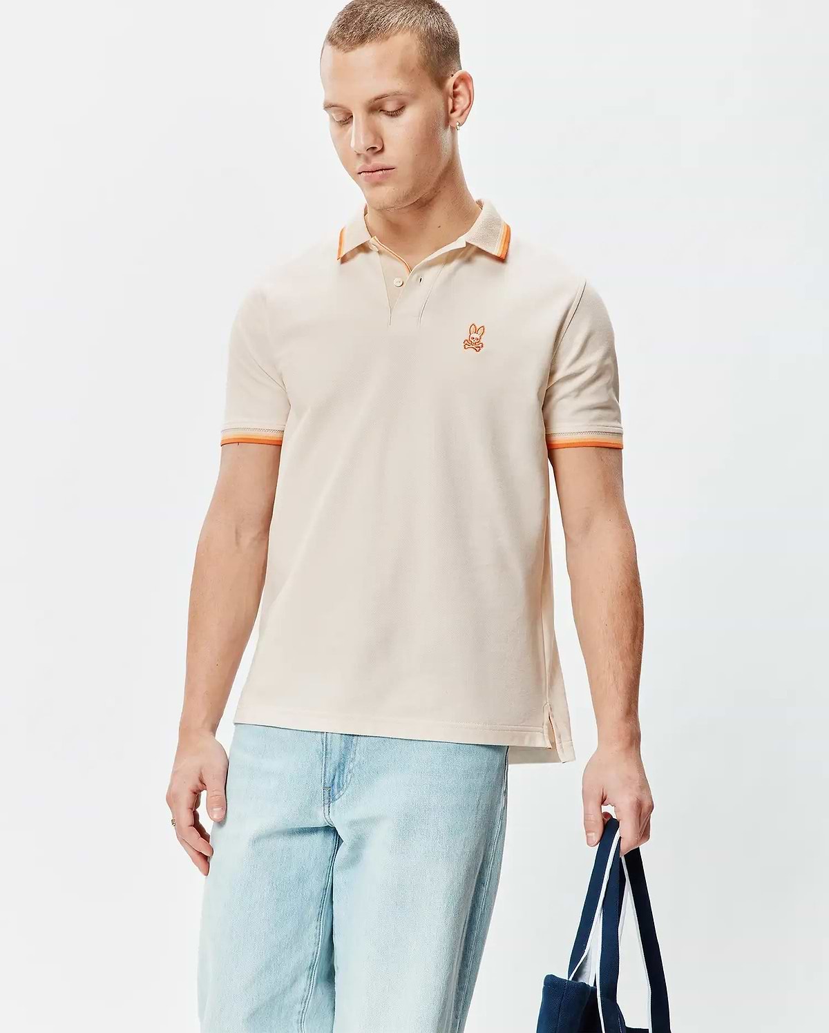 Mens Holden Pique Polo in classic fit, light beige pique with orange tipped collar and cuffs, featuring a small embroidered orange Psycho Bunny skull logo on the left chest.