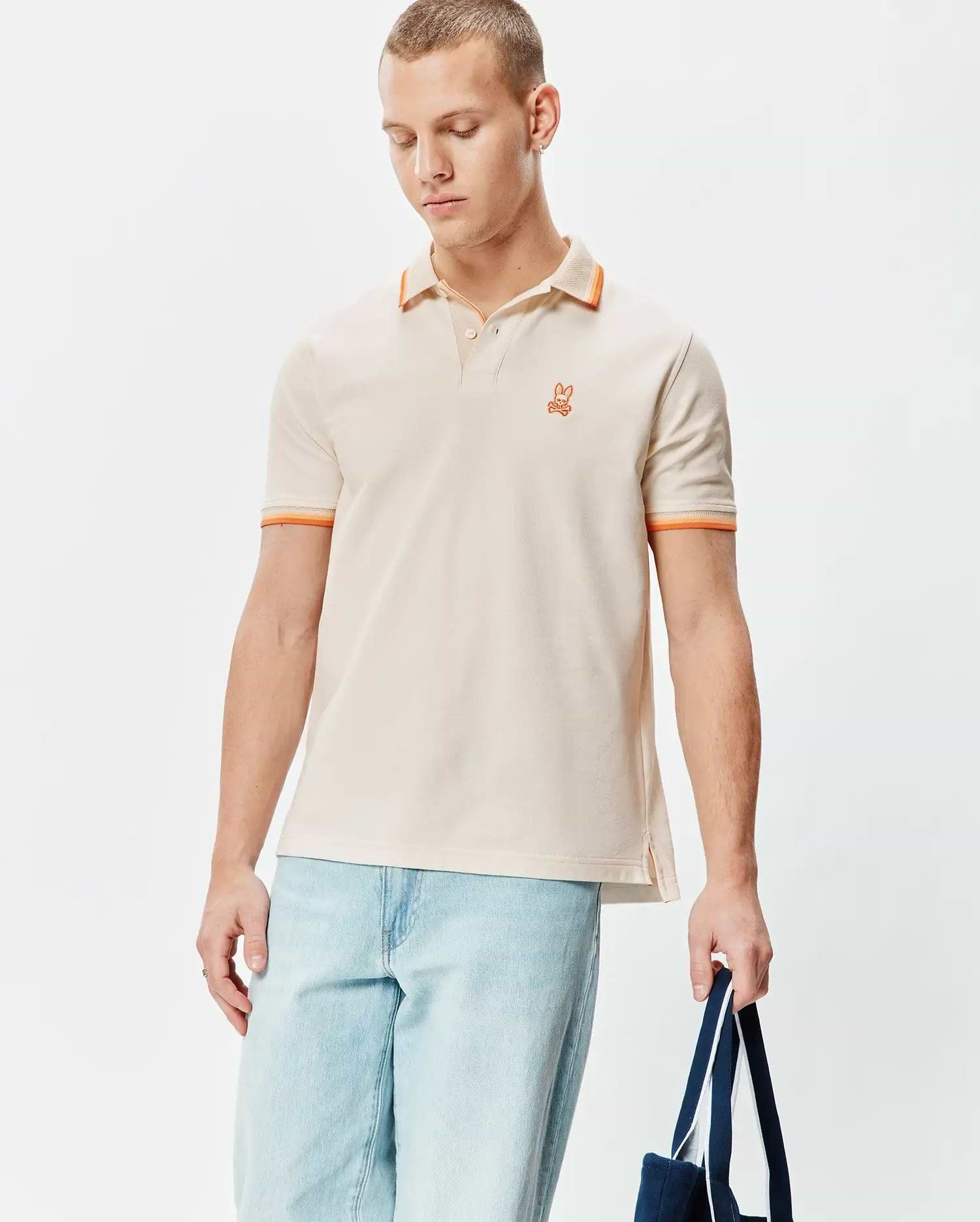 Mens Holden Pique Polo in classic fit, light beige pique with orange tipped collar and cuffs, featuring a small embroidered orange Psycho Bunny skull logo on the left chest.