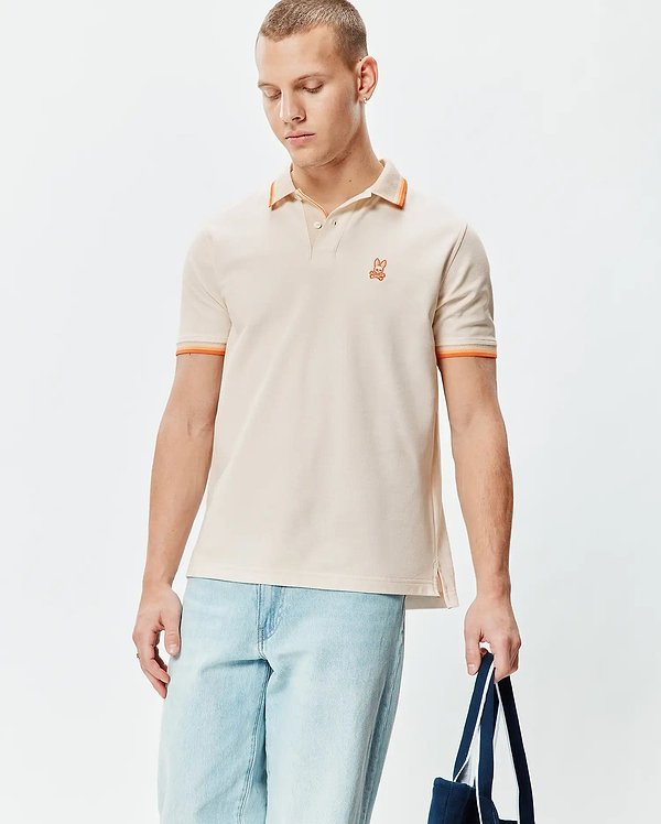 Mens Holden Pique Polo in classic fit, light beige pique with orange tipped collar and cuffs, featuring a small embroidered orange Psycho Bunny skull logo on the left chest.