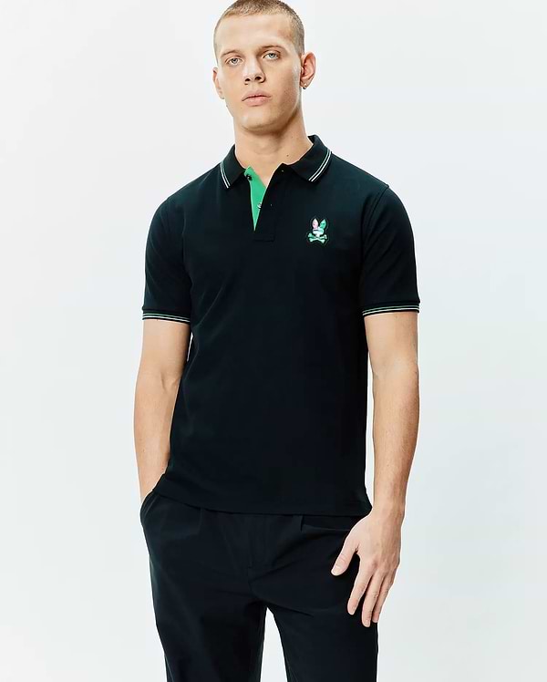 Mens Nate Pique Polo, a classic fit black pique polo with a vibrant green undercollar, white and green striped collar and cuffs, featuring an embroidered multi-color Psycho Bunny skull logo on the left chest.