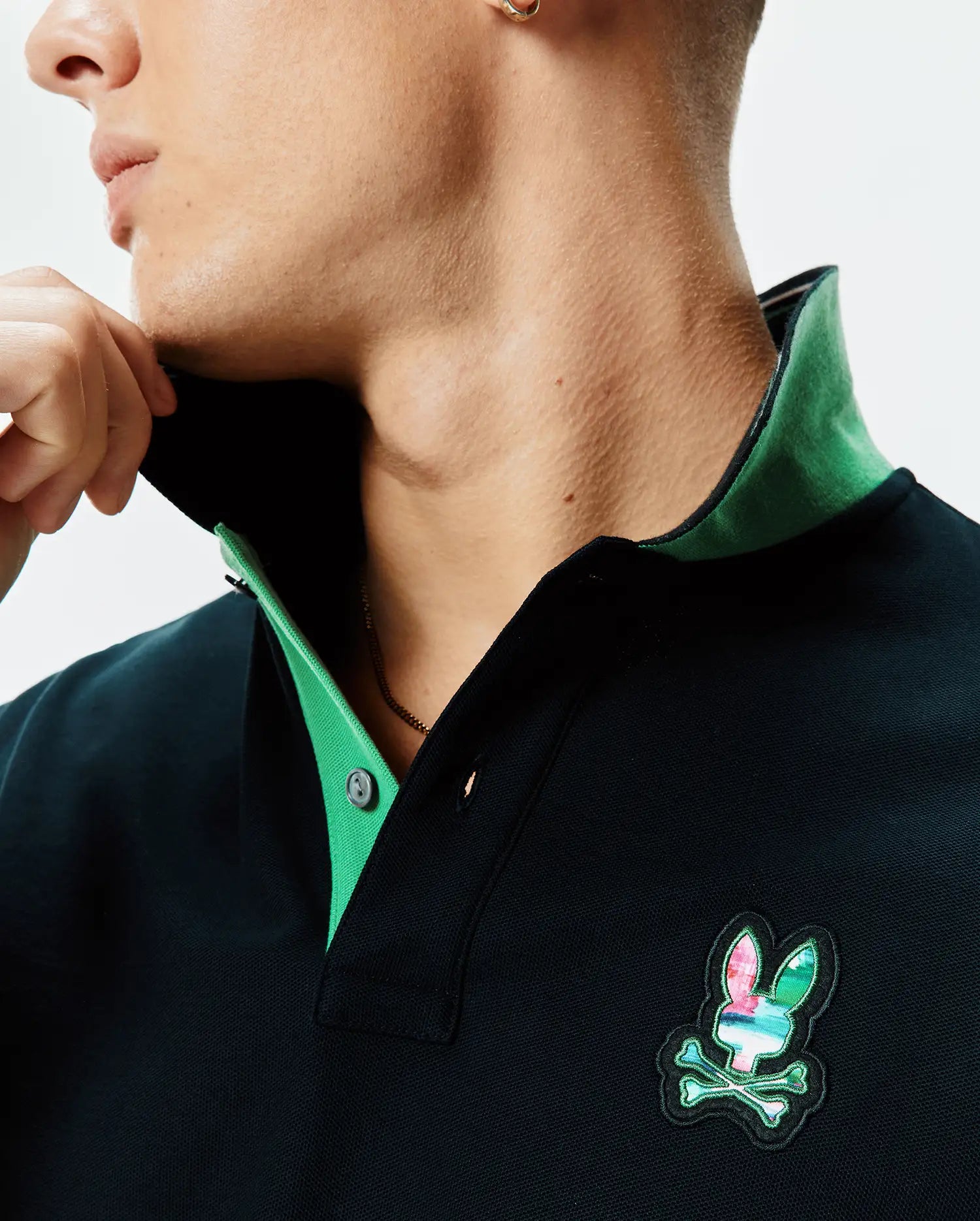 Mens Nate Pique Polo, a dark navy classic fit pique polo featuring a vibrant green contrast collar and a prominent multi-color embroidered Psycho Bunny skull logo on the chest.