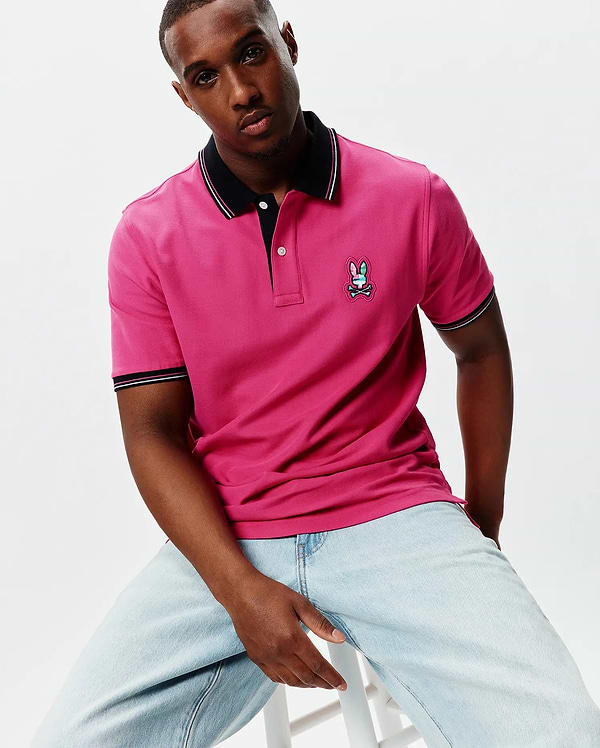 Mens Nate Pique Polo in vibrant fuchsia, featuring a classic fit, black ribbed collar and cuffs with white and pink tipping, and a colorful embroidered Psycho Bunny skull logo on the left chest.