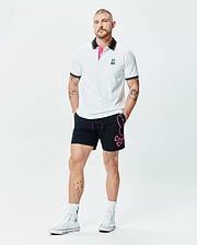 White Mens Nate Pique Polo with black collar, pink under-collar, striped cuffs, and embroidered Psycho Bunny logo, paired with black shorts featuring a large pink Psycho Bunny skull graphic.