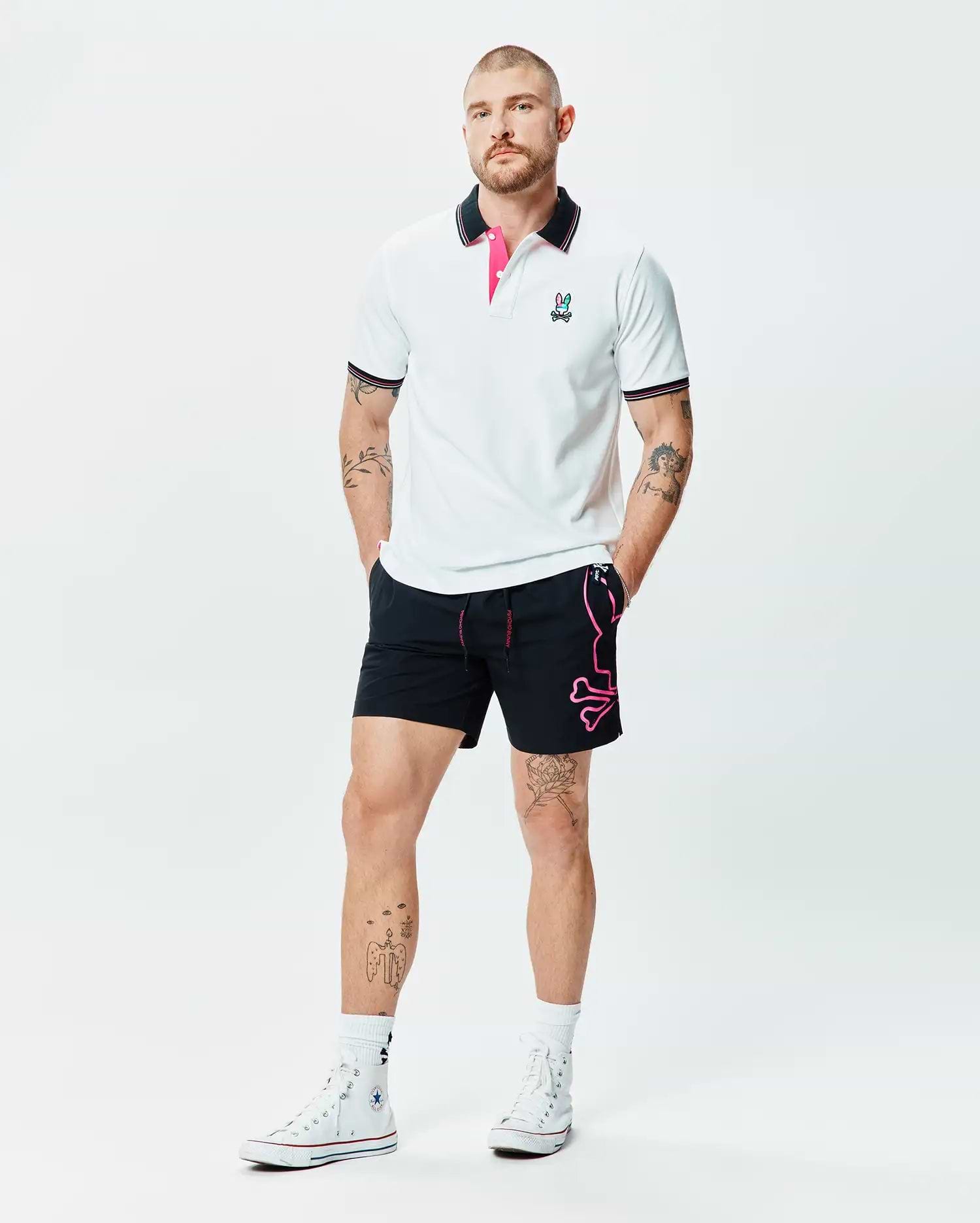 White Mens Nate Pique Polo with black collar, pink under-collar, striped cuffs, and embroidered Psycho Bunny logo, paired with black shorts featuring a large pink Psycho Bunny skull graphic.