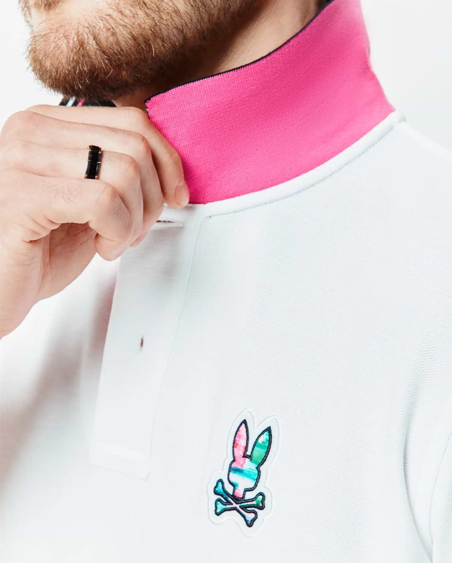 Mens Nate Pique Polo in white pique knit, featuring a vibrant pink contrast collar and a small, embroidered multicolor Psycho Bunny skull logo on the left chest.