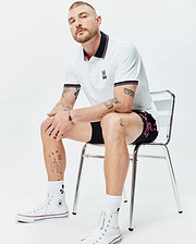 MENS NATE PIQUE POLO, a white classic pique polo with black and pink striped collar/cuffs, featuring a colorful embroidered Psycho Bunny logo. Black shorts with a vibrant pink Psycho Bunny side graphic.