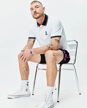 MENS NATE PIQUE POLO, a white classic pique polo with black and pink striped collar/cuffs, featuring a colorful embroidered Psycho Bunny logo. Black shorts with a vibrant pink Psycho Bunny side graphic.