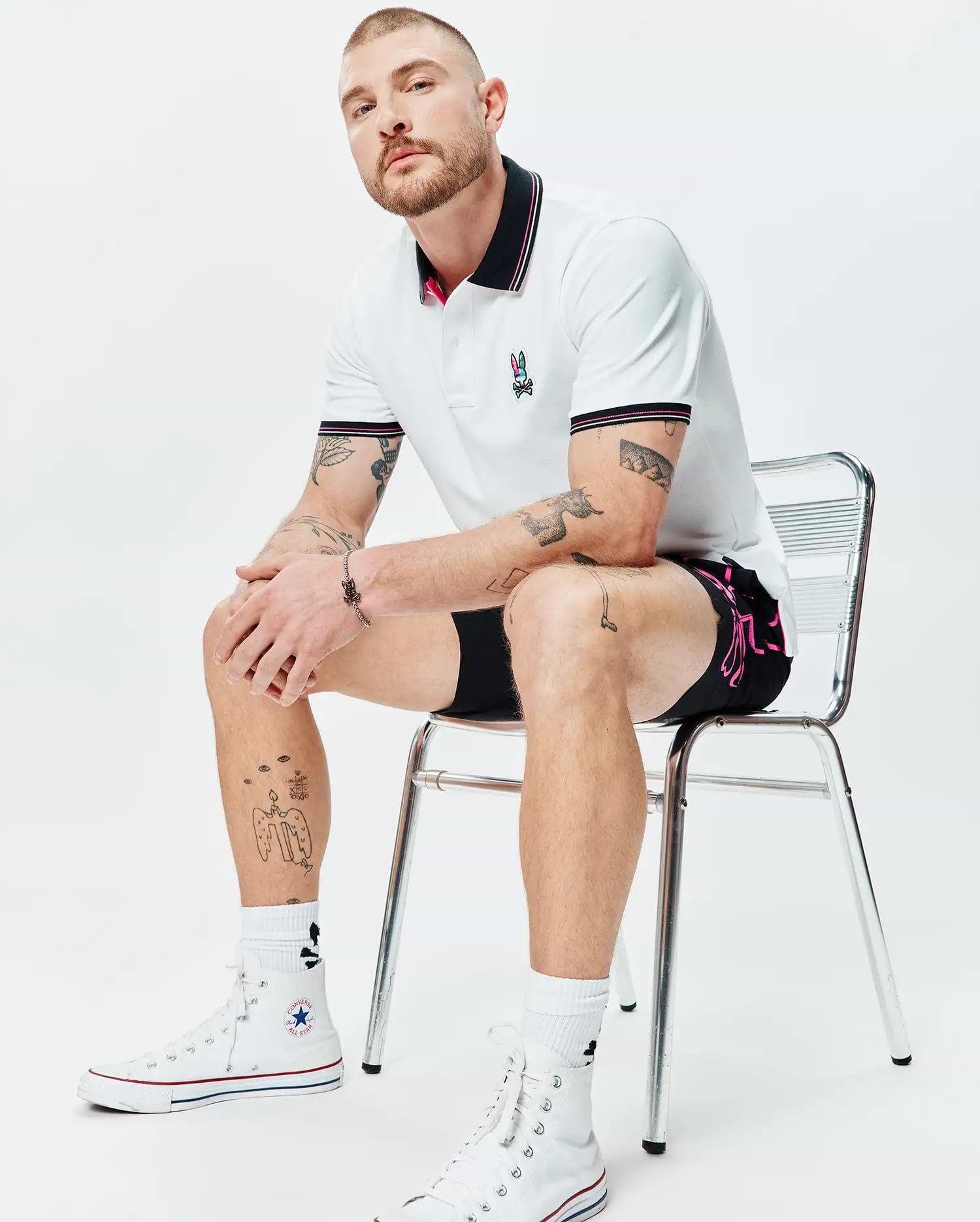 MENS NATE PIQUE POLO, a white classic pique polo with black and pink striped collar/cuffs, featuring a colorful embroidered Psycho Bunny logo. Black shorts with a vibrant pink Psycho Bunny side graphic.
