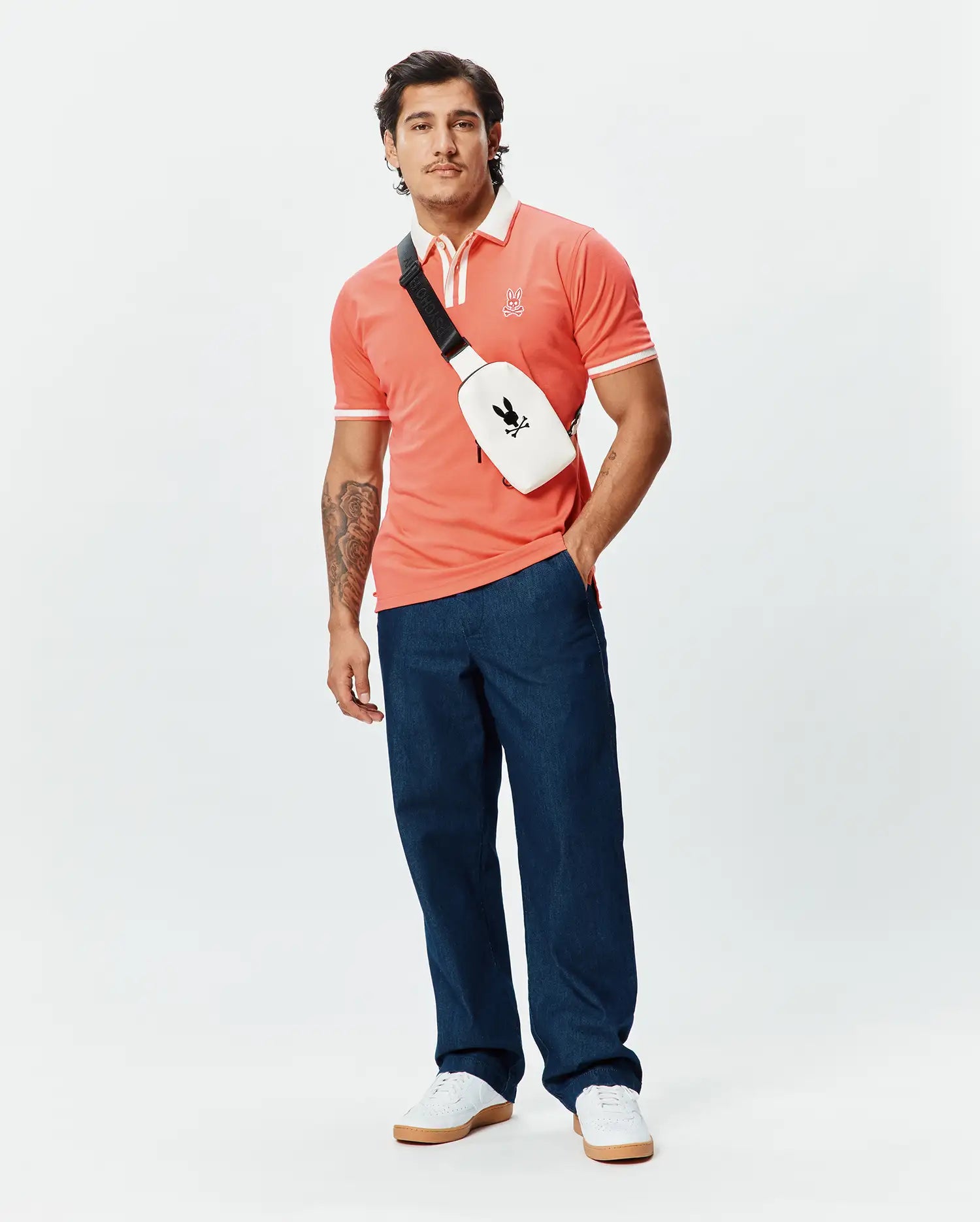 Mens Ridley Pique Polo in coral, featuring a classic fit, pique fabric, white contrast collar, white double-stripe ribbed cuffs, and a small embroidered Psycho Bunny skull logo on the left chest.