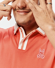 Mens Ridley Pique Polo, a classic fit coral pique polo with white striped collar and placket, featuring an embroidered white Psycho Bunny skull logo on the left chest.