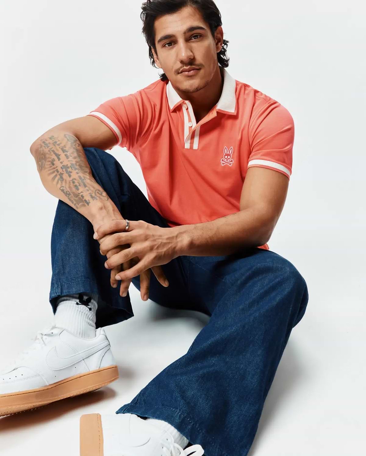 Mens Ridley Pique Polo in vibrant coral pique, classic fit, with a white ribbed collar, two-button placket, and white striped cuffs. Features a white embroidered Psycho Bunny skull logo on the left chest.