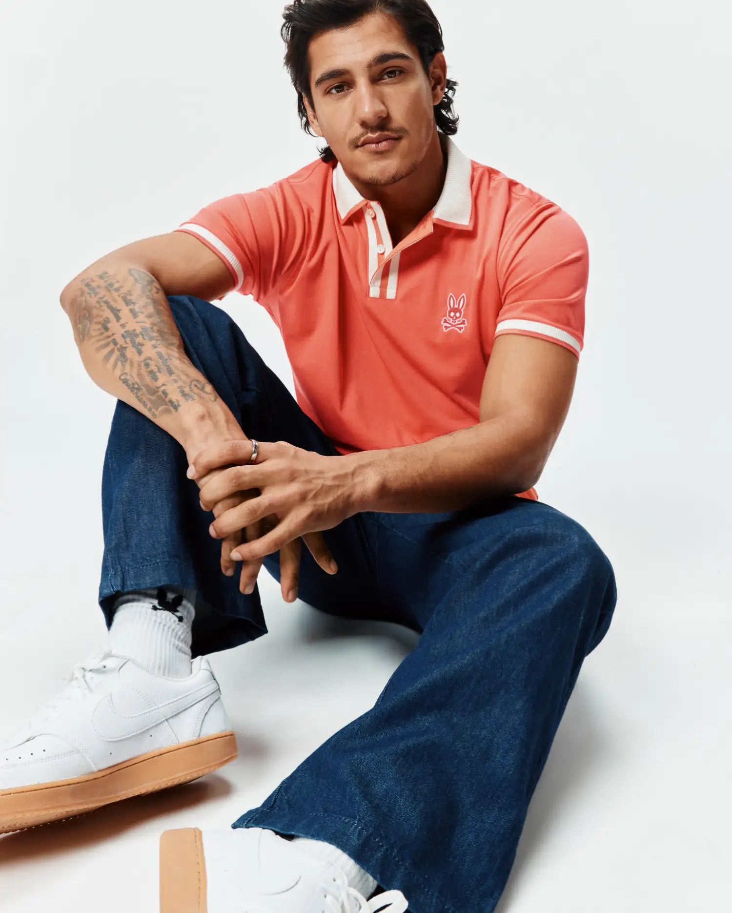 Mens Ridley Pique Polo in vibrant coral pique, classic fit, with a white ribbed collar, two-button placket, and white striped cuffs. Features a white embroidered Psycho Bunny skull logo on the left chest.