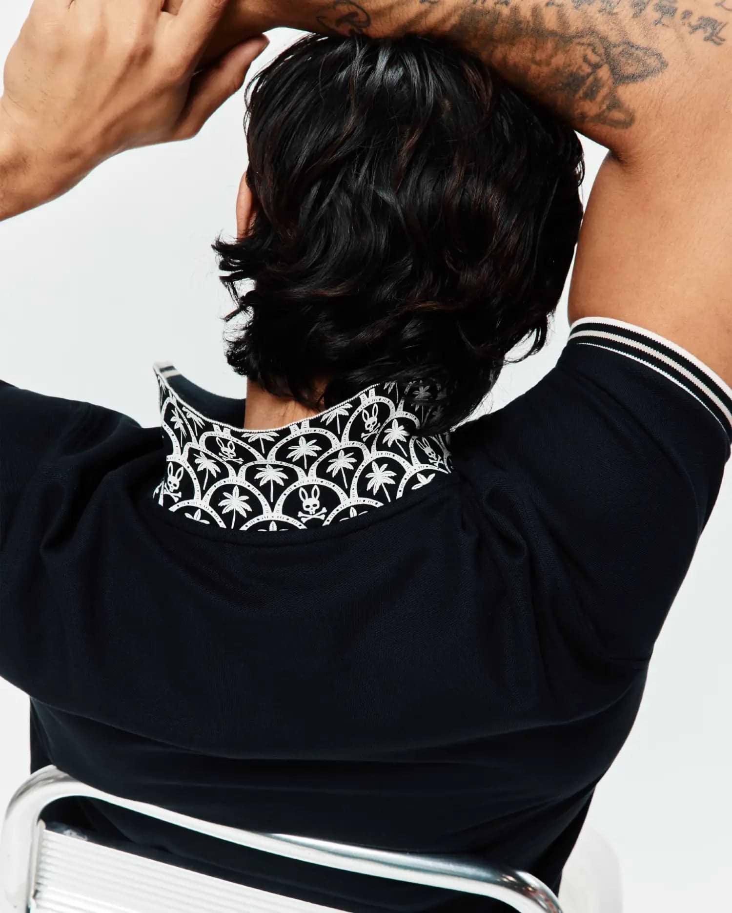 Black Mens Brad Pique Polo, classic pique knit, with a popped collar showcasing a white Psycho Bunny skull and palm tree pattern, plus white striped cuffs.