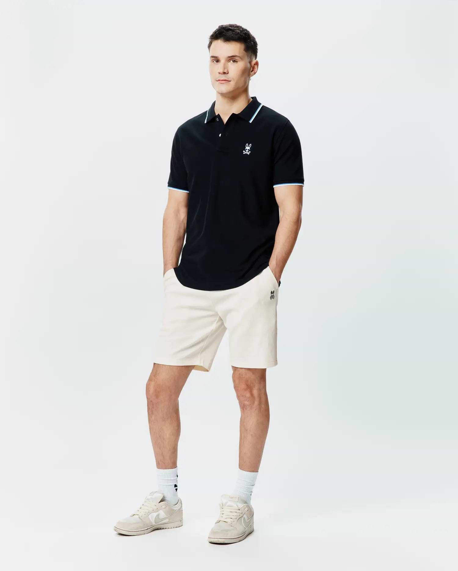 Black Mens Sunny Pique Polo, classic fit pique, with white and light blue striped collar and cuffs, featuring a small embroidered white Psycho Bunny skull logo on the left chest. Paired with cream ribbed shorts.