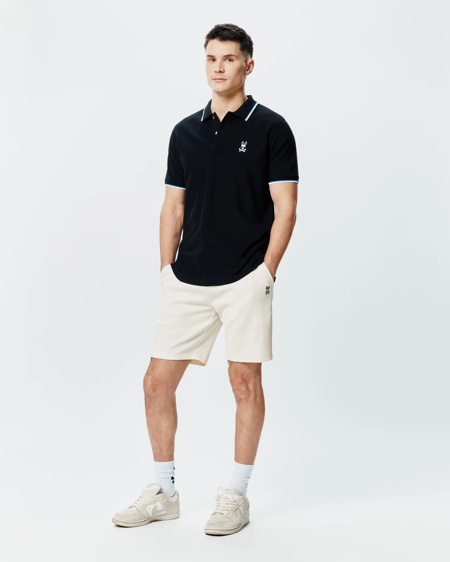 Black Mens Sunny Pique Polo, classic fit pique, with white and light blue striped collar and cuffs, featuring a small embroidered white Psycho Bunny skull logo on the left chest. Paired with cream ribbed shorts.