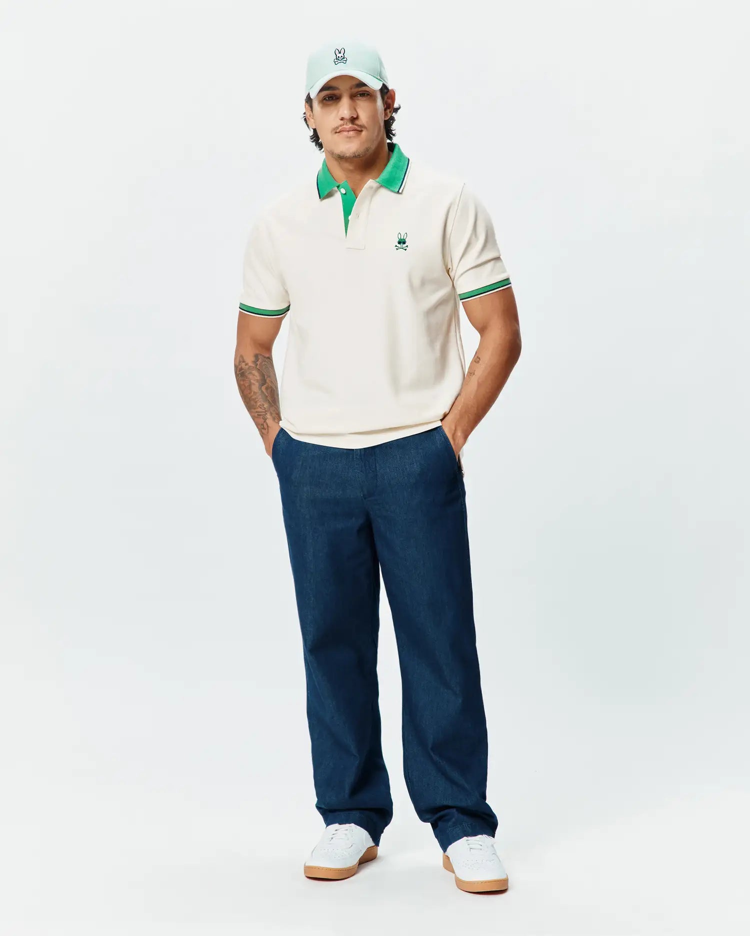 Mens Sunny Pique Polo, a classic fit cream pique polo shirt with a contrasting green collar, green and white striped cuffs, and a small embroidered green Psycho Bunny skull logo on the left chest.