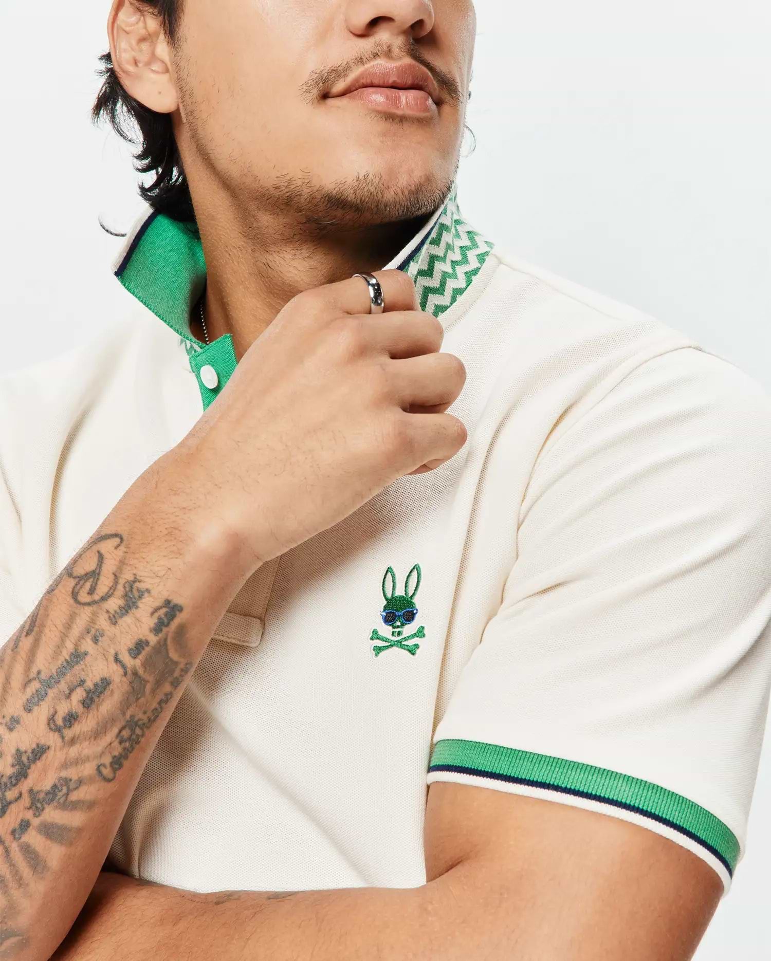Mens Sunny Pique Polo in cream pique, classic fit, featuring a green collar with white zig-zag pattern, striped green and navy cuffs, and an embroidered green bunny skull logo with blue sunglasses on the left chest.