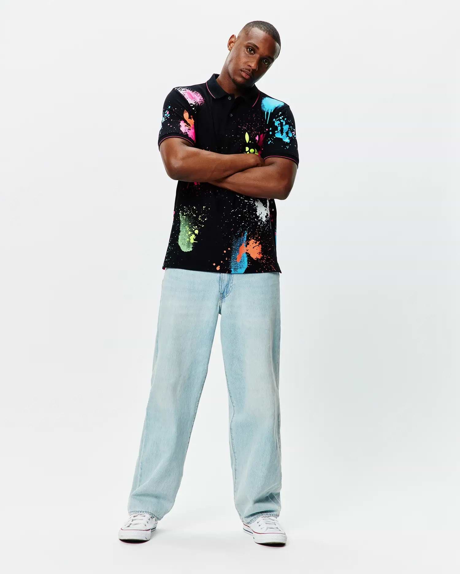 Quintin Aop Pique Polo: Black classic fit pique polo with vibrant all-over neon paint splatter print in pink, blue, green, orange, and white, featuring a small embroidered Psycho Bunny skull logo on the left chest.