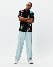 Quintin Aop Pique Polo: Black classic fit pique polo with vibrant all-over neon paint splatter print in pink, blue, green, orange, and white, featuring a small embroidered Psycho Bunny skull logo on the left chest.
