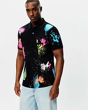 Mens Quintin Aop Pique Polo, a black classic fit pique polo with an all-over vibrant neon paint splatter print in pink, blue, green, orange, and white, featuring subtle Psycho Bunny skull logos and pink trim.