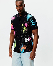 Mens Quintin Aop Pique Polo, a black classic fit pique polo with an all-over vibrant neon paint splatter print in pink, blue, green, orange, and white, featuring subtle Psycho Bunny skull logos and pink trim.