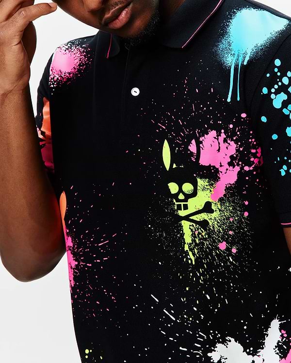Mens Quintin Aop Pique Polo, a black pique polo with a vibrant all-over neon paint splatter print in pink, lime, blue, and orange, featuring a large central Psycho Bunny skull logo graphic.