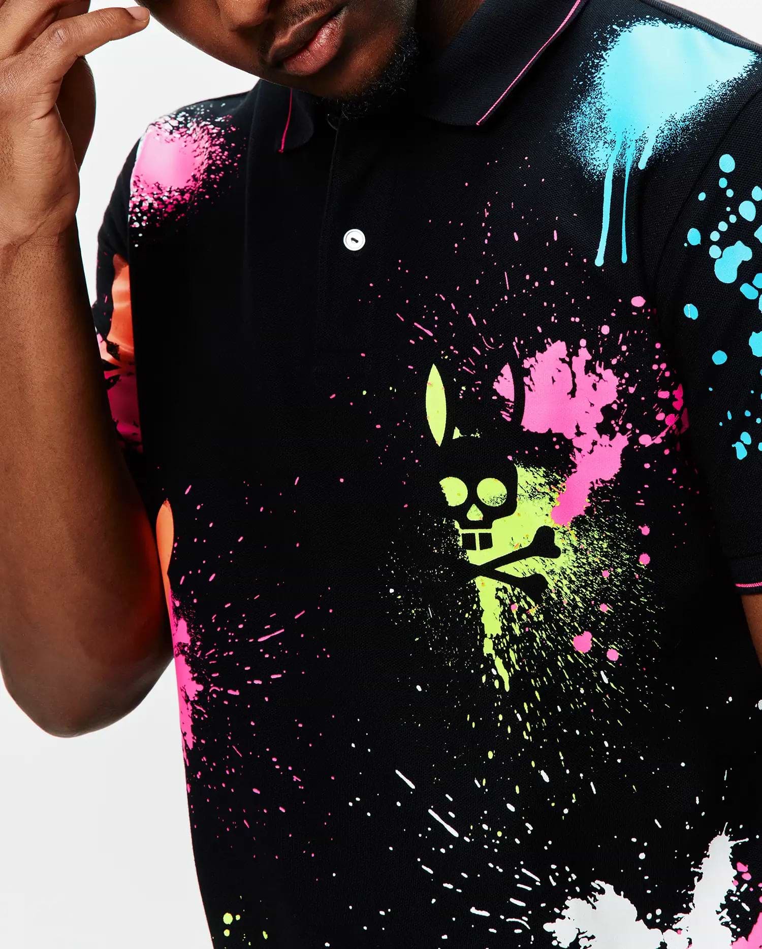 Mens Quintin Aop Pique Polo, a black pique polo with a vibrant all-over neon paint splatter print in pink, lime, blue, and orange, featuring a large central Psycho Bunny skull logo graphic.