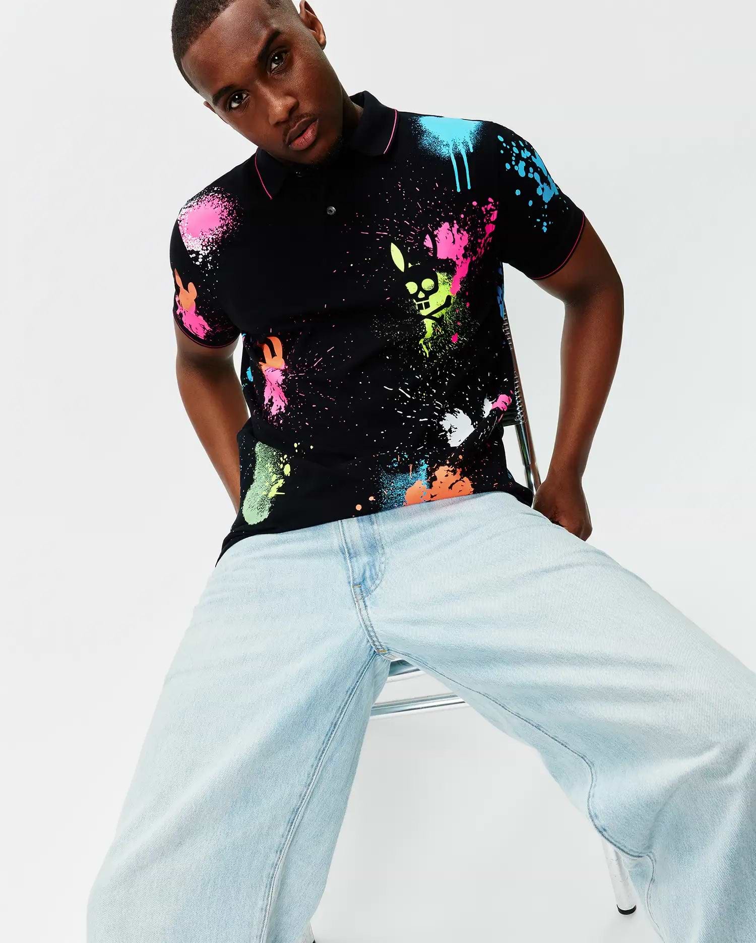 Mens Quintin Aop Pique Polo, a black classic fit pique polo with vibrant neon pink, blue, green, orange, and white paint splatter print, featuring the Psycho Bunny skull logo integrated into the design and pink trim.