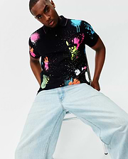 Mens Quintin Aop Pique Polo, a black classic fit pique polo with vibrant neon pink, blue, green, orange, and white paint splatter print, featuring the Psycho Bunny skull logo integrated into the design and pink trim.