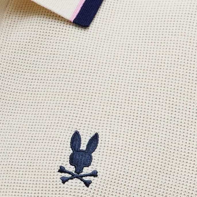 Mens Dax Knit Polo in cream textured knit, featuring a navy and pink striped ribbed collar and a small navy embroidered Psycho Bunny skull logo on the chest.