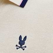 Mens Dax Knit Polo in cream textured knit, featuring a navy and pink striped ribbed collar and a small navy embroidered Psycho Bunny skull logo on the chest.