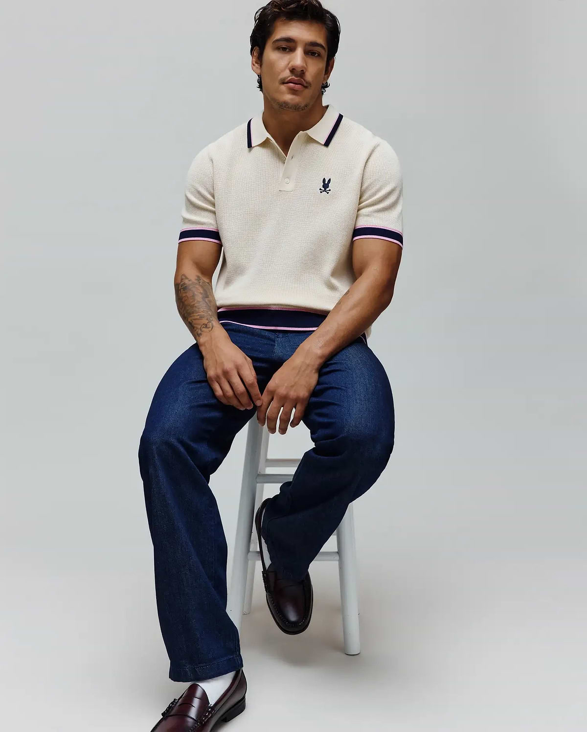 Mens Dax Knit Polo in cream with a classic fit, featuring a fine knit texture, navy and pink striped collar, sleeve cuffs, and hem, and a small embroidered Psycho Bunny skull logo on the left chest.