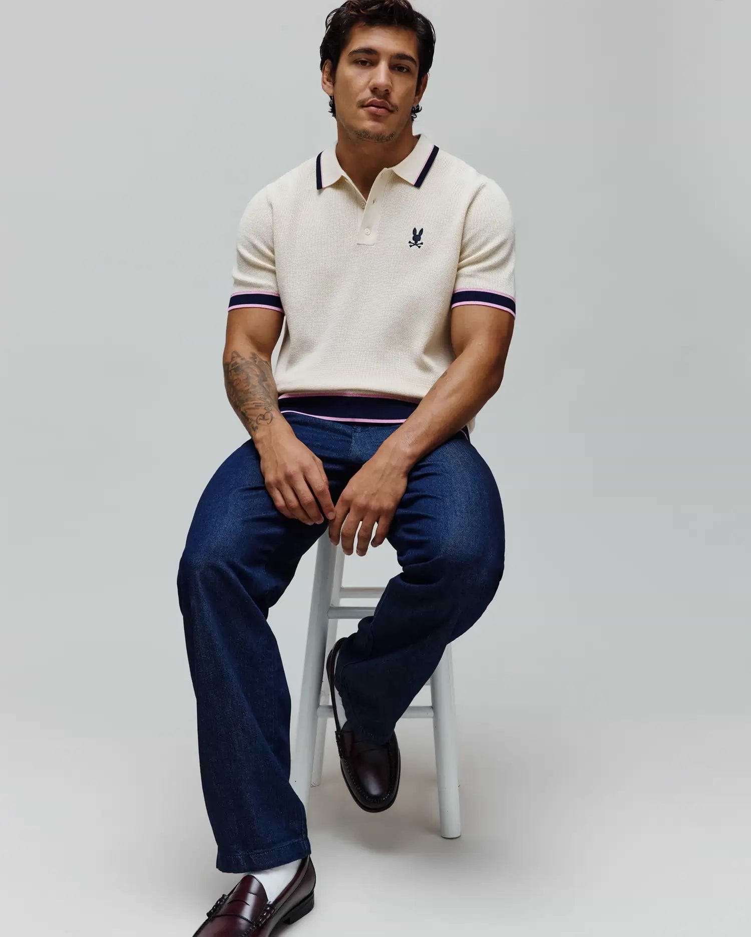 Mens Dax Knit Polo in cream with a classic fit, featuring a fine knit texture, navy and pink striped collar, sleeve cuffs, and hem, and a small embroidered Psycho Bunny skull logo on the left chest.